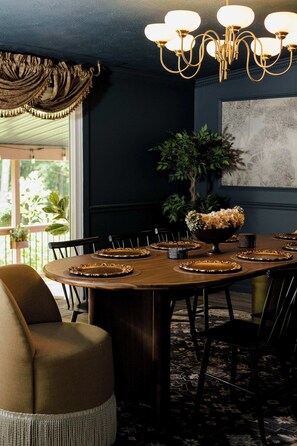 A dining area with a round table set for a meal, surrounded by chairs, with a chandelier above and a large plant in the background.