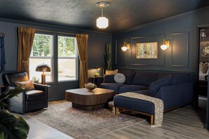 A modern living room with a dark blue sofa, a leather armchair, and a coffee table. There are two windows with curtains, a floor lamp, and wall-mounted lights.