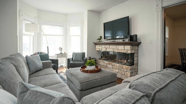 A living room with a grey sectional sofa, a stone fireplace, a flat-screen TV, and a dining area with chairs. - Heated Pool and Hot Tub near Downtown Lake Geneva! (Lake Geneva)