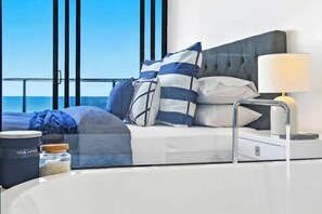 A modern hotel room with a bed, a nightstand, a lamp, a window with a view of the ocean, and a small table with a candle and a jar. - Luxe by the Beach at Oracle Level 13 (Gold Coast City)