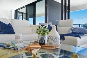 A modern living room with a glass coffee table, a white sofa, and a view of the sky through large windows. - Luxe by the Beach at Oracle Level 13 (Gold Coast City)
