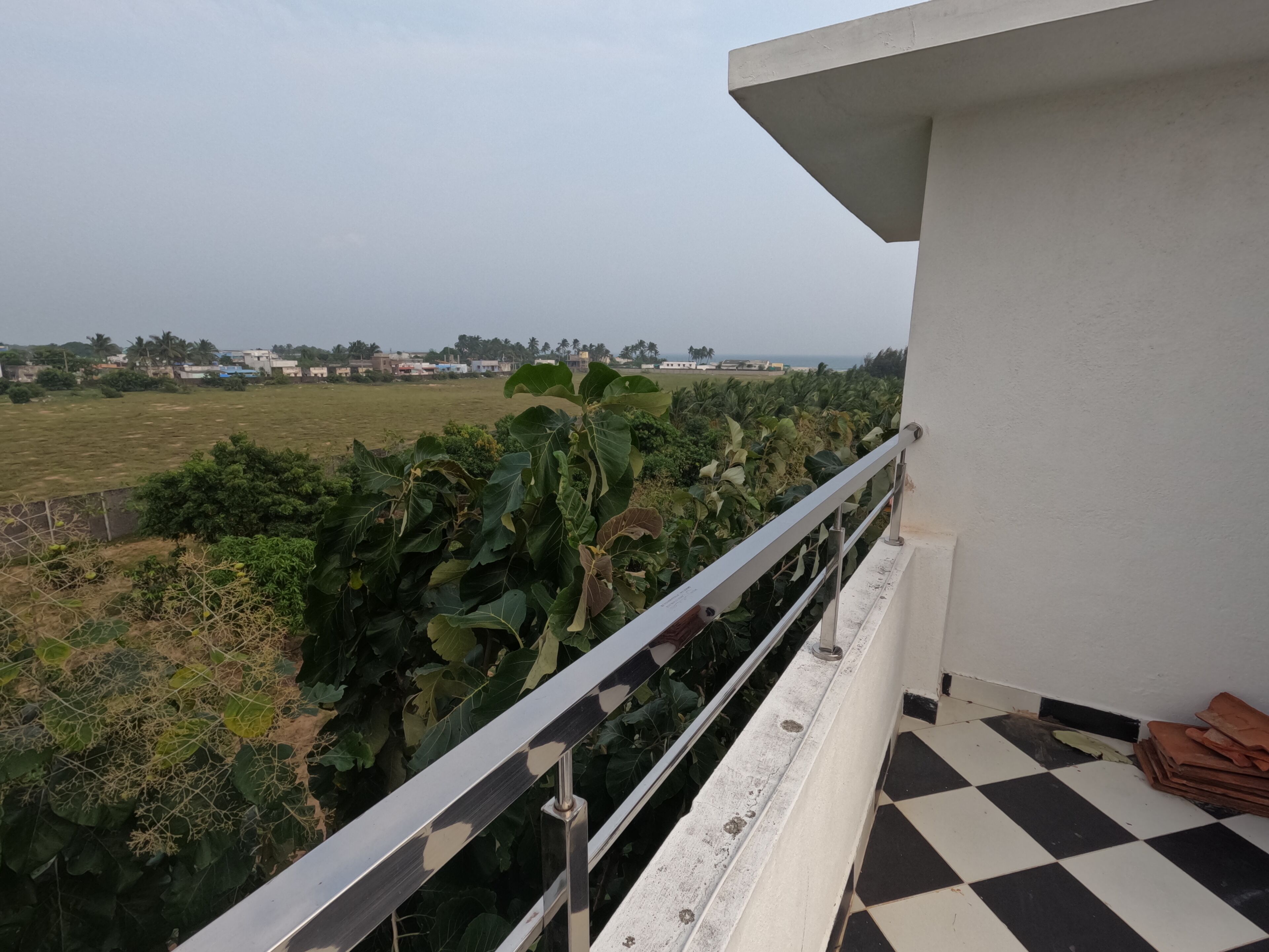 Ein Balkon mit Metallgeländer, einem Schachbrettboden und Blick auf ein grünes Feld mit Gebäuden in der Ferne.