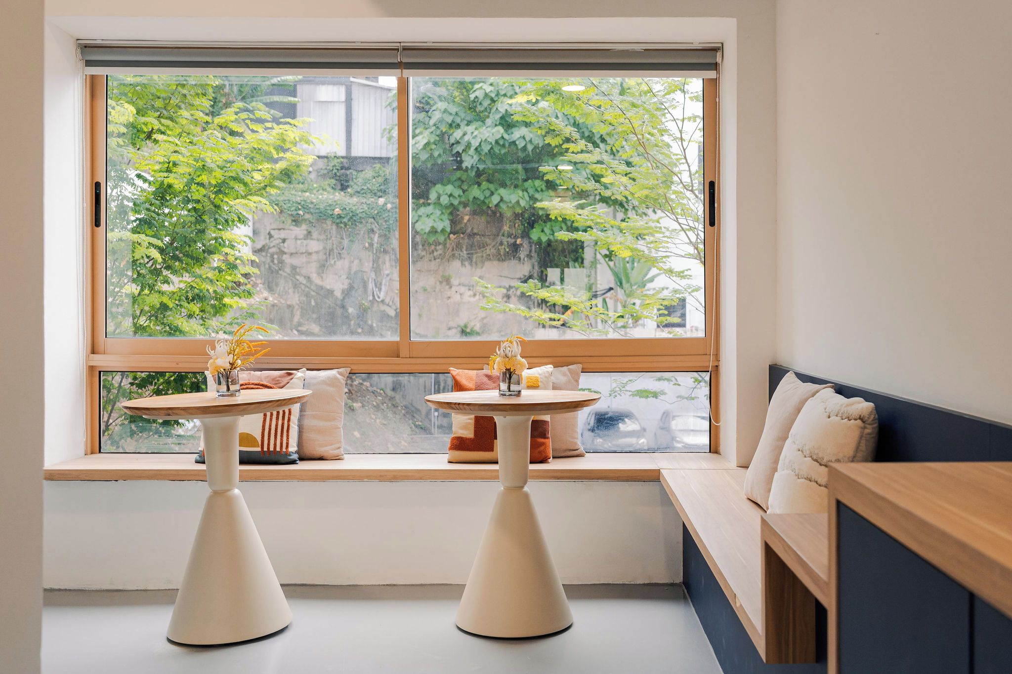 Una habitación con un ventanal grande, dos mesas blancas sobre pedestal, un banco y un banco de madera a lo largo de la pared.