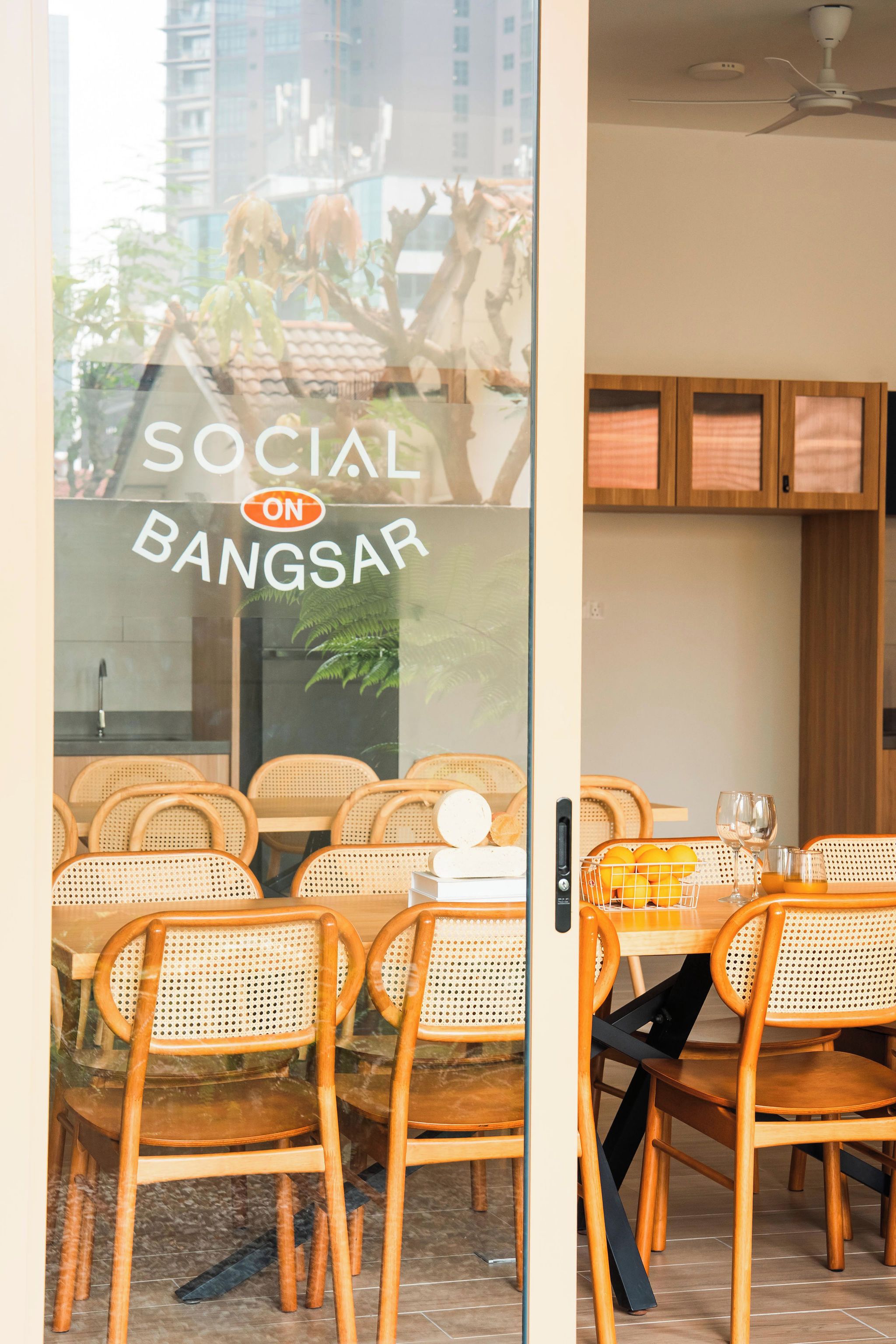 A restaurant interior with wicker chairs, a dining table, and a view of the city through a glass door.