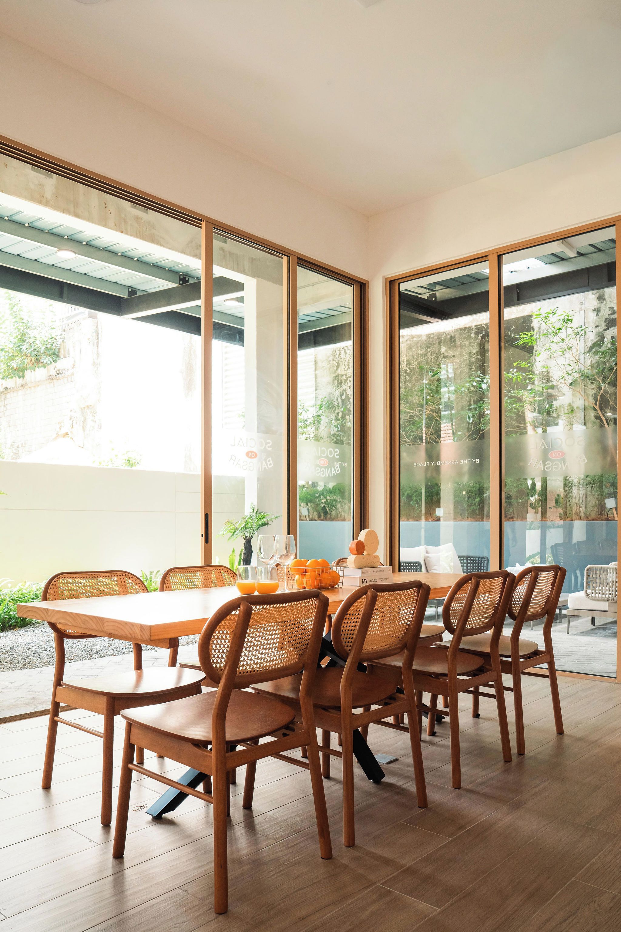 A dining area with a table set for six, surrounded by wicker chairs, and large windows offering a view of greenery outside.