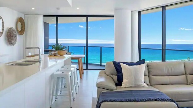 A modern living room with a large window overlooking the sea, a sofa, and a kitchen area with bar stools. - Beach Haven at Oracle Level 14 (Gold Coast City)