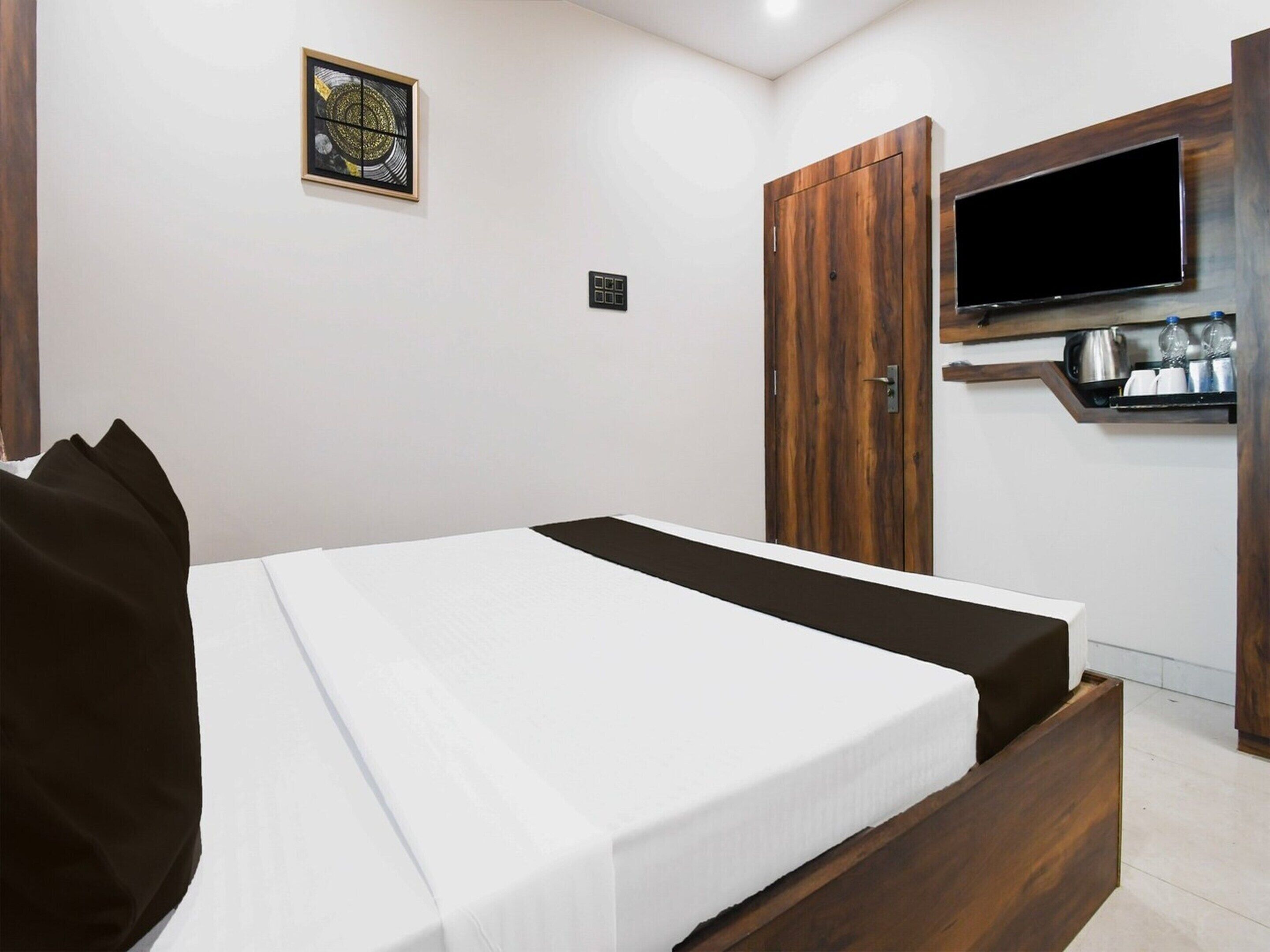 A bedroom with a bed, a wooden wardrobe, a television mounted on the wall, and a small shelf with items.