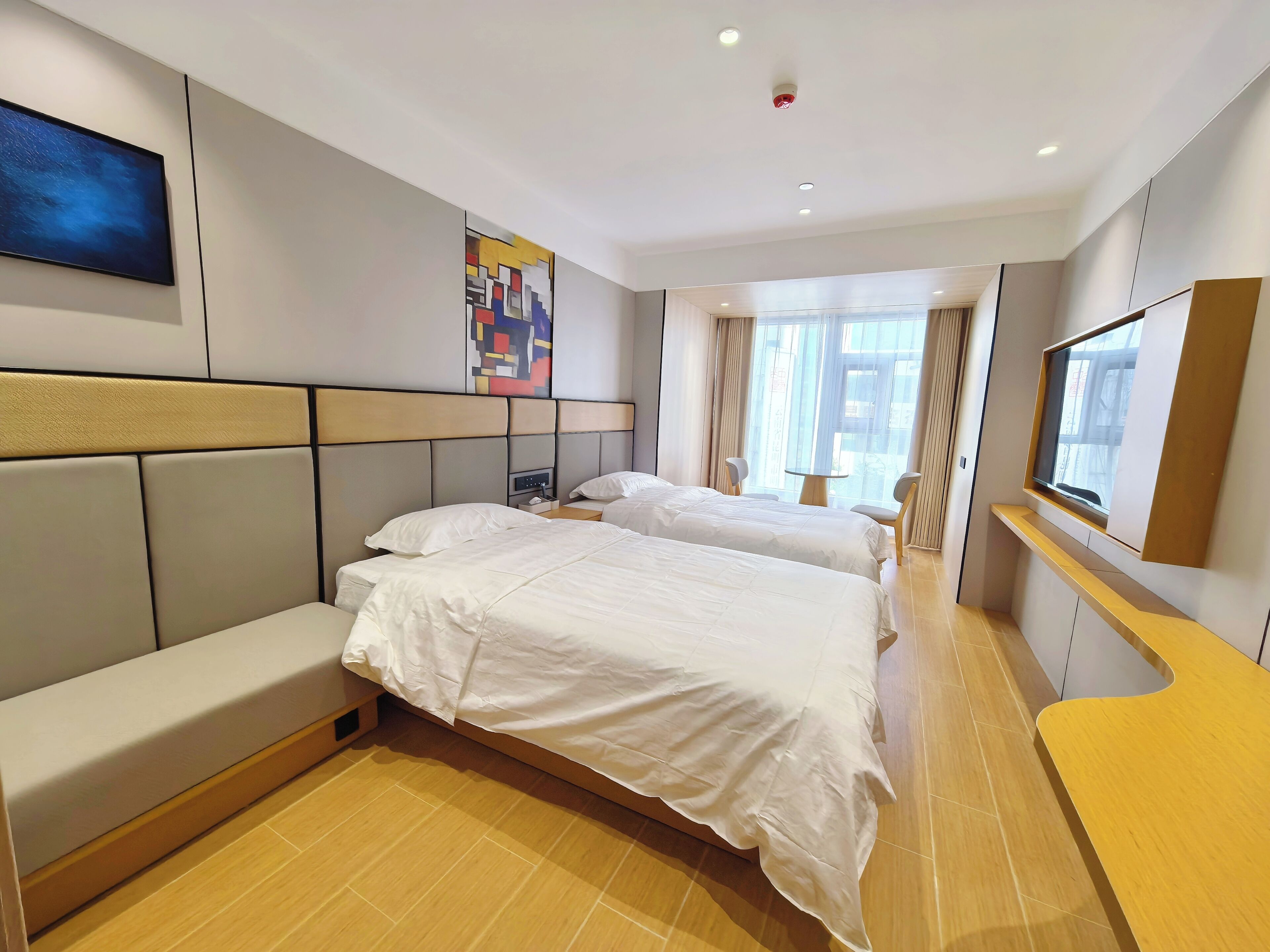 Quarto de hotel com duas camas, área de estar, televisão, obra de arte na parede e janela com cortinas.