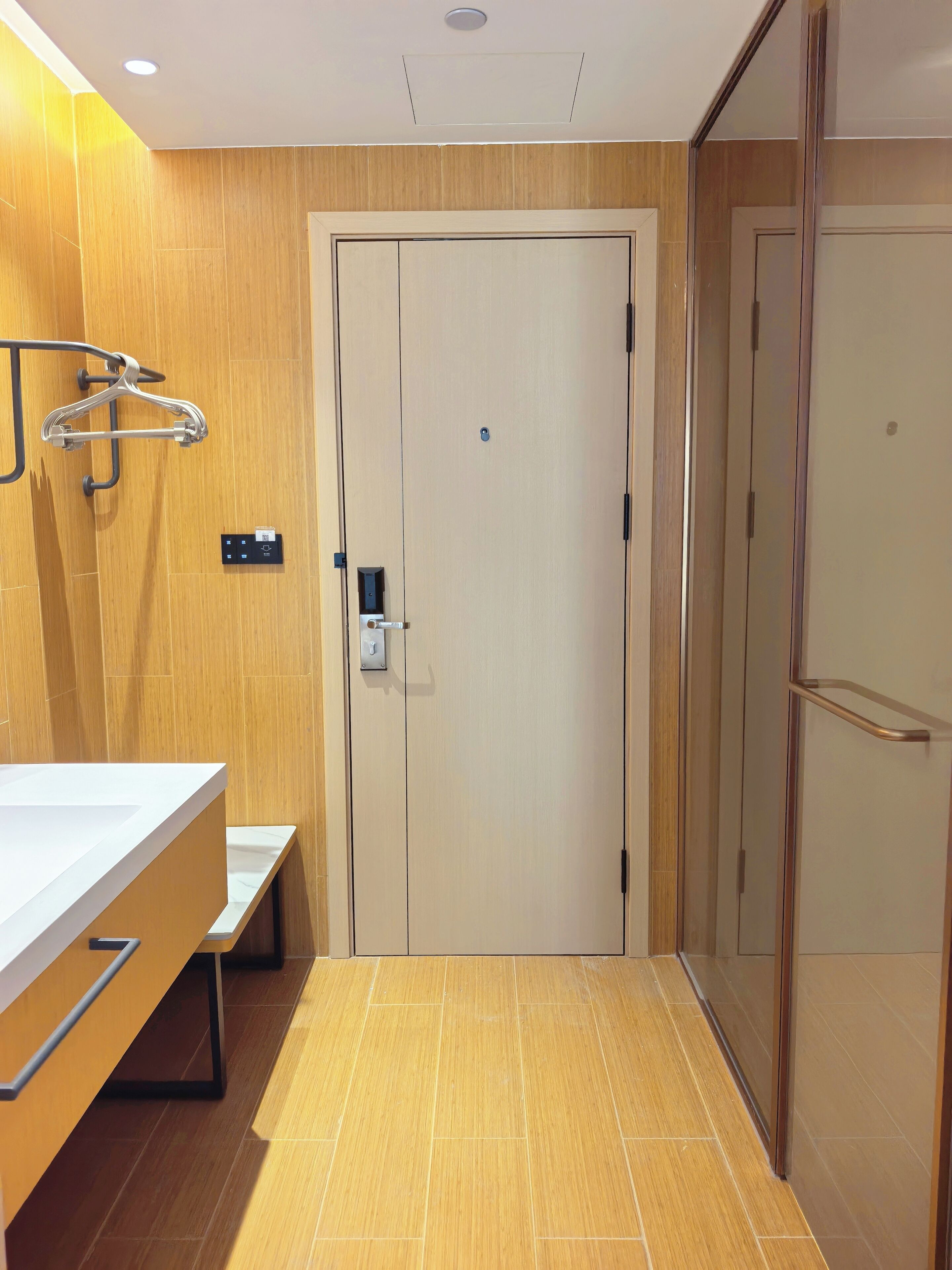 A hotel room with a wooden floor, a white door, a bathroom with a mirror and towel rack, and a small bench.