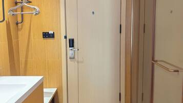 A hotel room with a wooden floor, a white door, a bathroom with a mirror and towel rack, and a small bench.