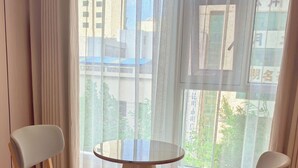 A hotel room with a round wooden table, two grey upholstered chairs, a bed with white bedding, and large windows with a view of buildings and greenery. - Kunming Fish Inn City Center Nanping Street Branch (Kunming)