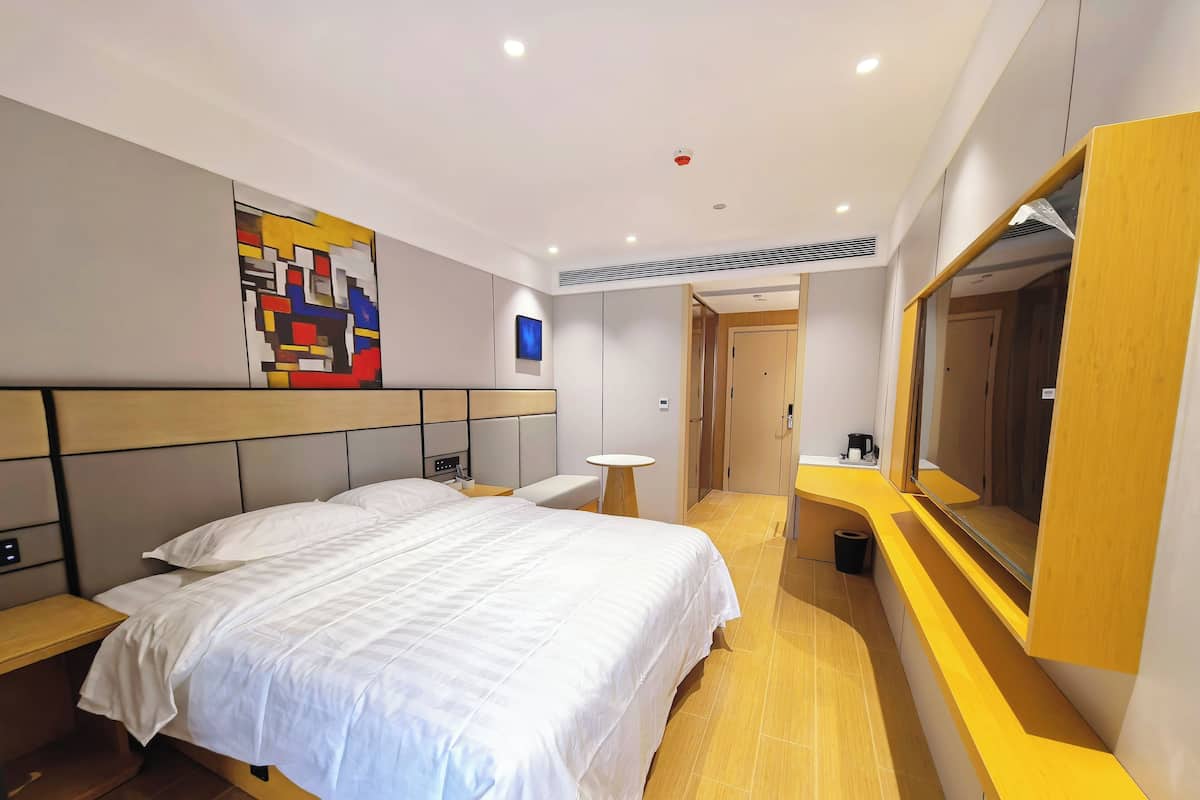 A modern hotel room with a large bed, bedside tables, a small round table, a mirror, and a door leading to another room.