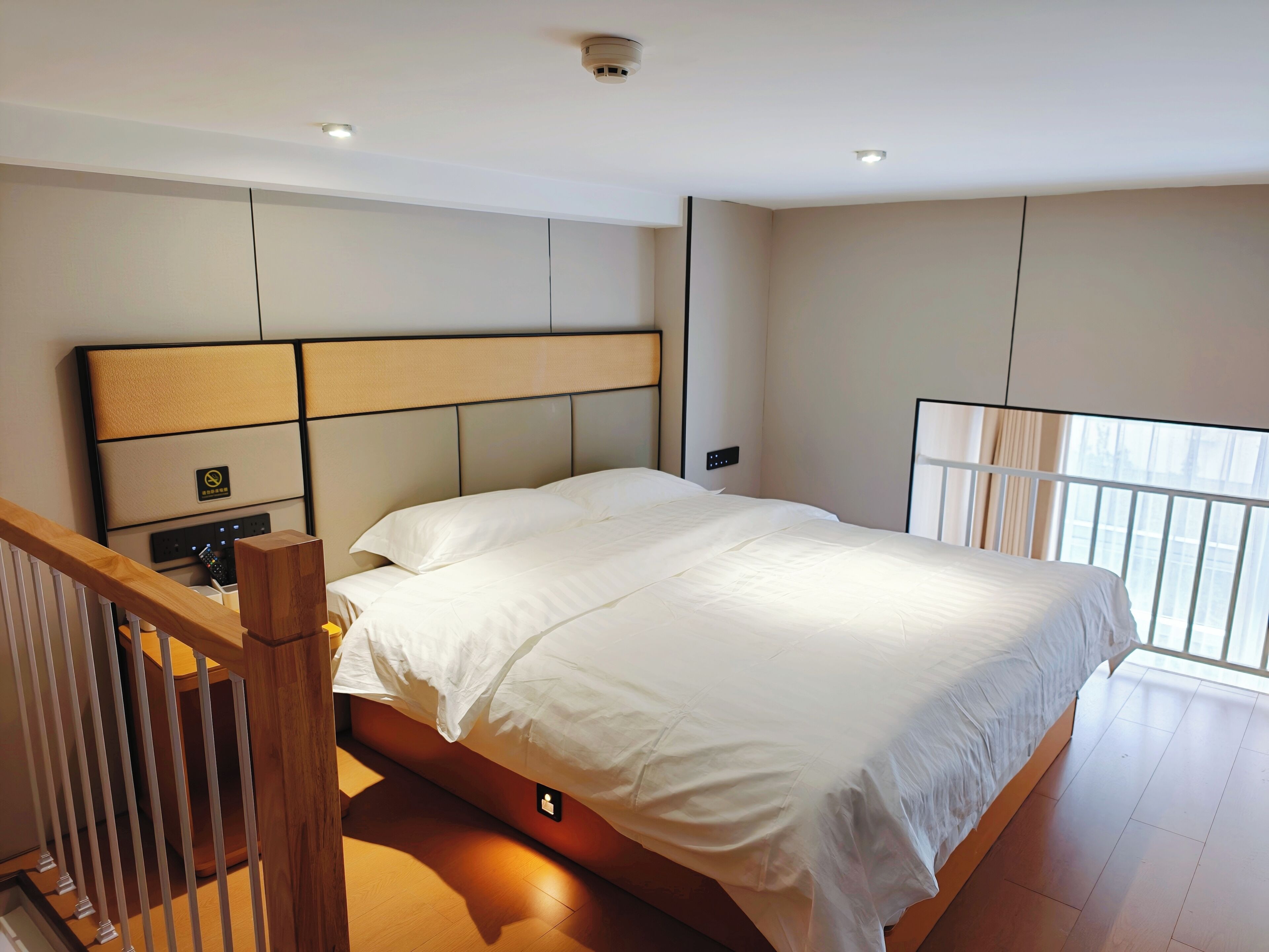Une chambre d’hôtel moderne dotée d’un grand lit, d’une tête de lit avec un panneau de commande, d’un escalier en bois et d’une fenêtre offrant une vue.