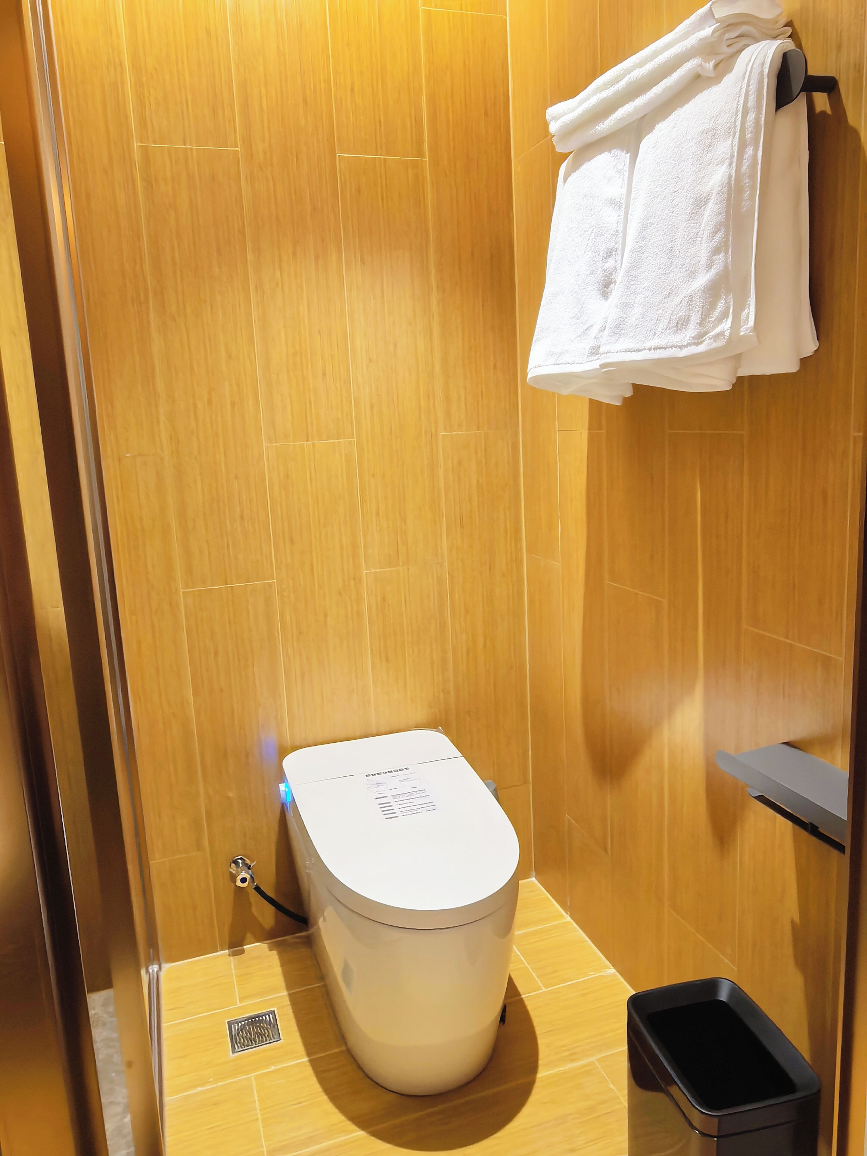 A small, compact bathroom with a white toilet, a black trash bin, and a towel rack with a white towel.