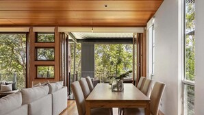 Un comedor con mesa y sillas de madera, un sofá y ventanas amplias con vistas a árboles.