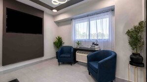 Une chambre moderne équipée d’un téléviseur à écran plat, de deux fauteuils bleus, d’une petite table avec une plante et d’une fenêtre ornée de rideaux légers.