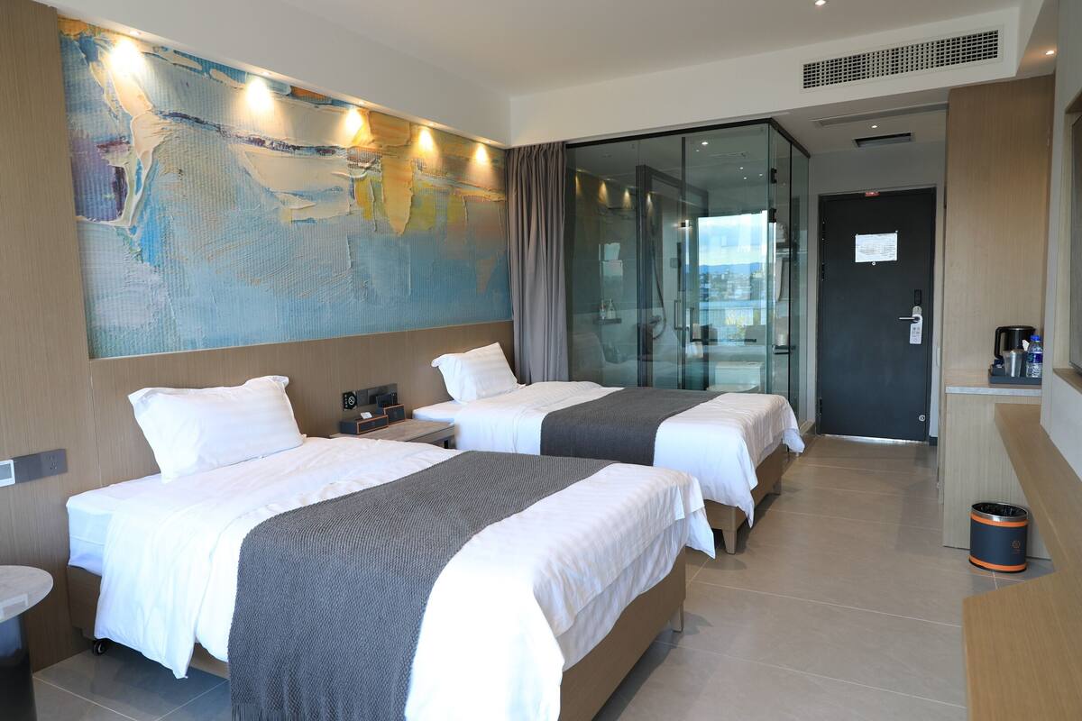 A hotel room with two beds, a large abstract painting on the wall, a glass door leading to a balcony, and a small kitchenette area.