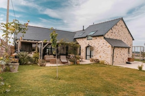 A stone house with a sloped roof, a covered patio with outdoor furniture, and a small garden area.