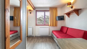 Ein kleines Zimmer mit einem roten Sofa, einem kleinen Tisch, einem an der Wand montierten Fernseher und einem Fenster mit Vorhängen.