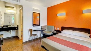A hotel room with a bed, a desk, a chair, a small table, a mirror, and a bathroom with a sink and toilet. - Queen Bed|Nsmk|Thornton (Thornton)
