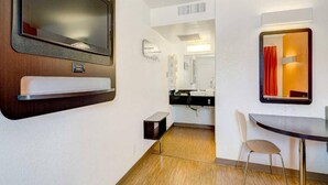 A compact hotel room with a bed, a small desk, a chair, a TV, a mirror, and a bathroom with a sink and mirror. - Queen Bed|Nsmk|Thornton (Thornton)