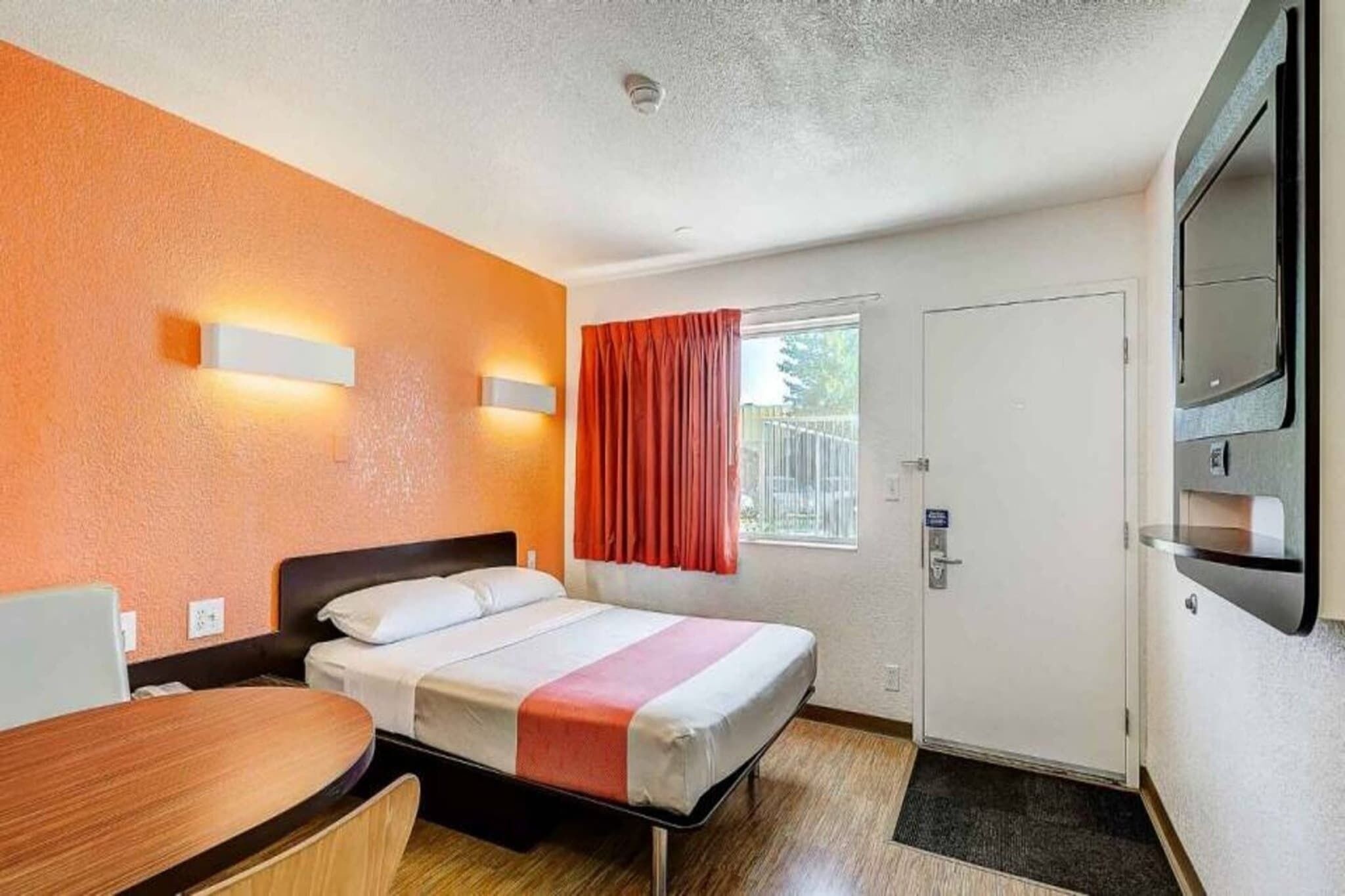 A hotel room with a bed, a desk, a TV, a window with curtains, and a door.