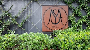 A sign with a logo and the word 'MYSA' against a dark wooden background, surrounded by greenery.