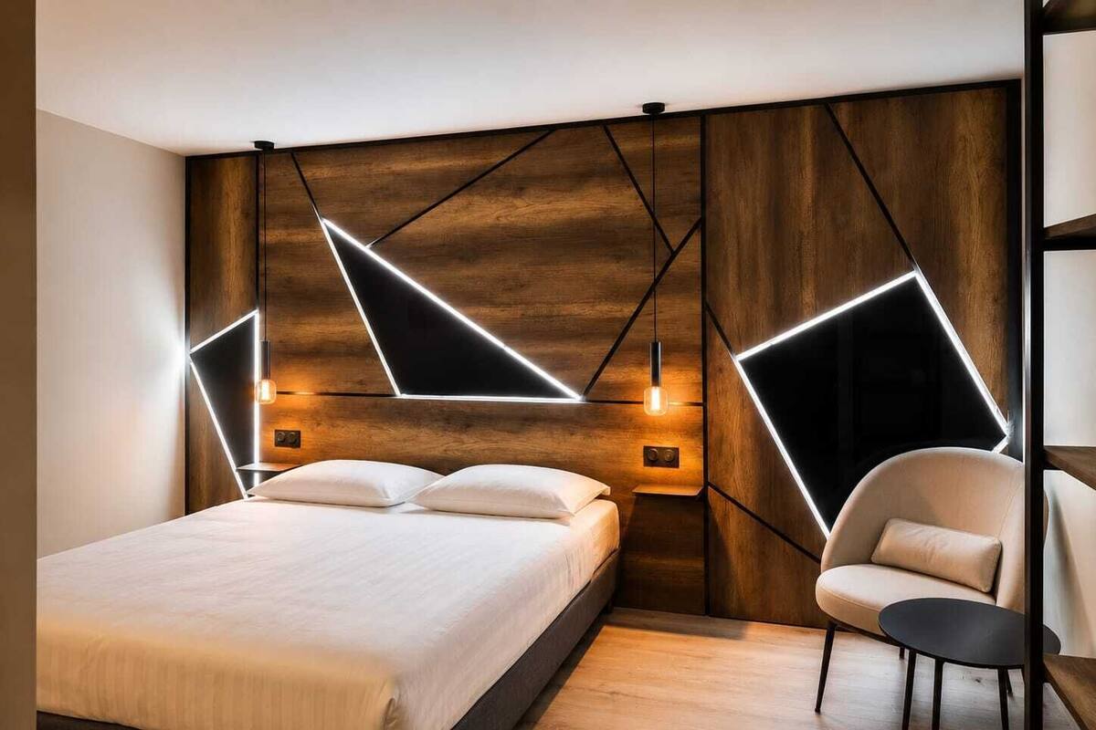 Un dormitorio moderno con cama, mesitas de noche, una silla e iluminación geométrica en la pared.