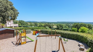 Um parquinho com balanços e estruturas de escalada, cercado por um arbusto e com vista para colinas ao longe.