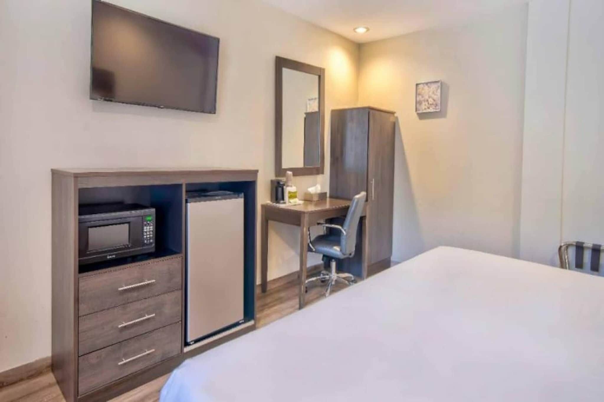 Habitación de hotel con cama, escritorio con silla, microondas, armario y televisor de pantalla plana montado en la pared.
