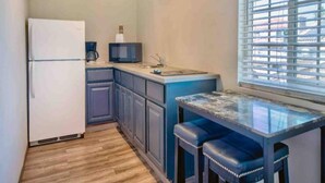 A compact kitchen with a refrigerator, microwave, and a small sink. There is a window with blinds and a wooden floor.