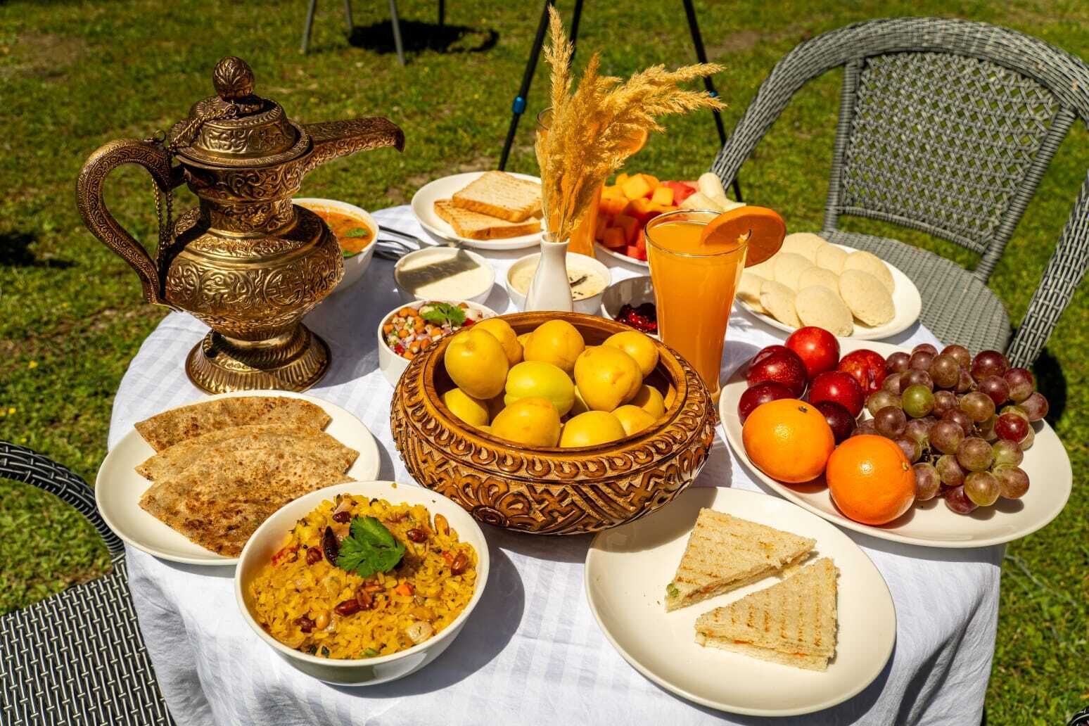 Una mesa al aire libre con diversas comidas, como pan, arroz, frutas y una jarra de bronce decorativa.