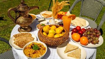 Una mesa al aire libre con diversas comidas, como pan, arroz, frutas y una jarra de bronce decorativa.