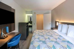 A hotel room with a bed, a desk, a chair, a TV, a minibar, and a bathroom door.