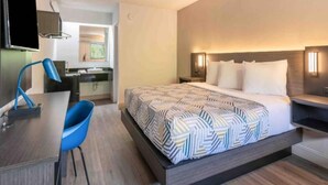 A hotel room with a bed, bedside table, chair, and a small desk with a chair. - Clean ADA King Bed|Nsmk (Charlotte NC)