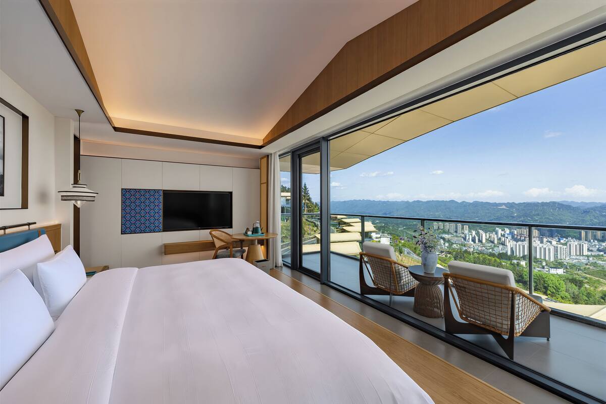 A modern hotel room with a large bed, a TV, a balcony with wicker chairs, and a city view.