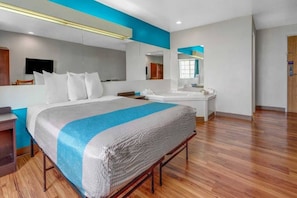 A modern hotel room with a large bed, a bathroom with a mirror, and a wooden floor. - 1 Queen Bed | Non-Smoking (Idaho Falls)