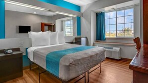 Ein modernes Hotelzimmer mit Bett, Nachttisch, Fernseher, Fenster mit Vorhängen und Schreibtisch mit Stuhl.