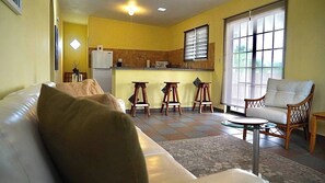 Ein Wohnzimmer mit einer Couch, einem Sessel, einem kleinen Tisch und einer Küchenzeile mit Kühlschrank, Mikrowelle und Sitzhocker.