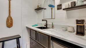 Un baño con lavamanos, espejo, repisas con tazas y una cafetera.