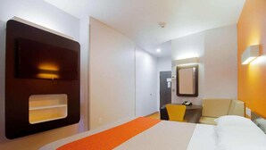 Habitación de hotel con una cama, un escritorio, una silla, un espejo y un televisor montado en la pared.