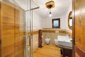 Un baño con suelo de madera, cabina de ducha de vidrio, bidé, lavabo y tocador de madera.
