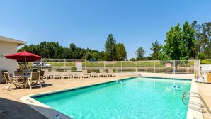 A rectangular swimming pool with a tiled edge, surrounded by a white fence. There are lounge chairs and a red umbrella on the poolside.
