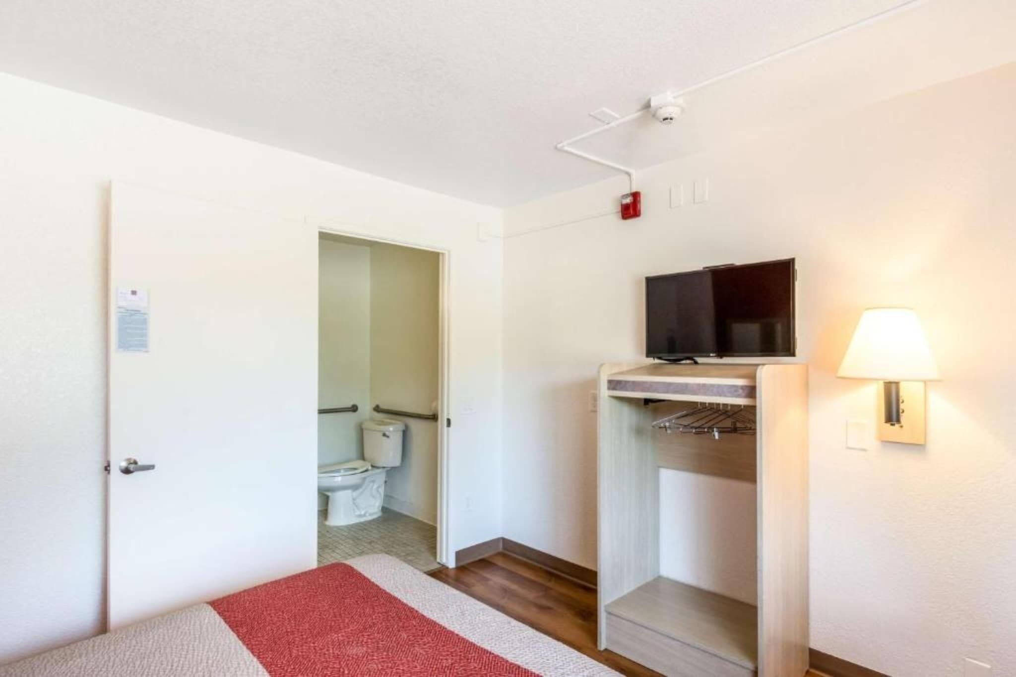 A hotel room with a bed, a TV, a lamp, a shelf, and a door leading to a bathroom.