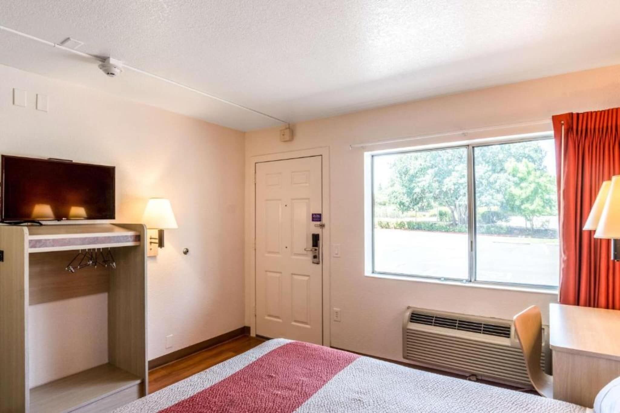 Une chambre d’hôtel avec un lit, un bureau avec une télévision, une fenêtre avec des rideaux et une porte.