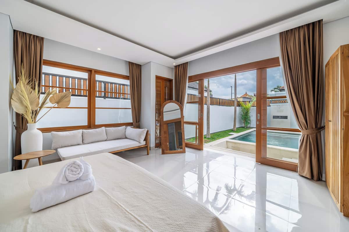 A hotel room with a large bed, a sofa, a small table, a vase with dried plants, and a view of a swimming pool.
