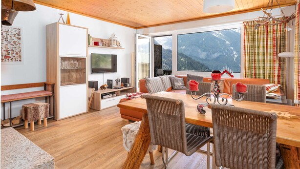 A cozy living room with a wooden ceiling, a large window with a mountain view, a dining table with wicker chairs, and a television on a white cabinet. - Mountain View Apartment in Grindelwald (Grindelwald)