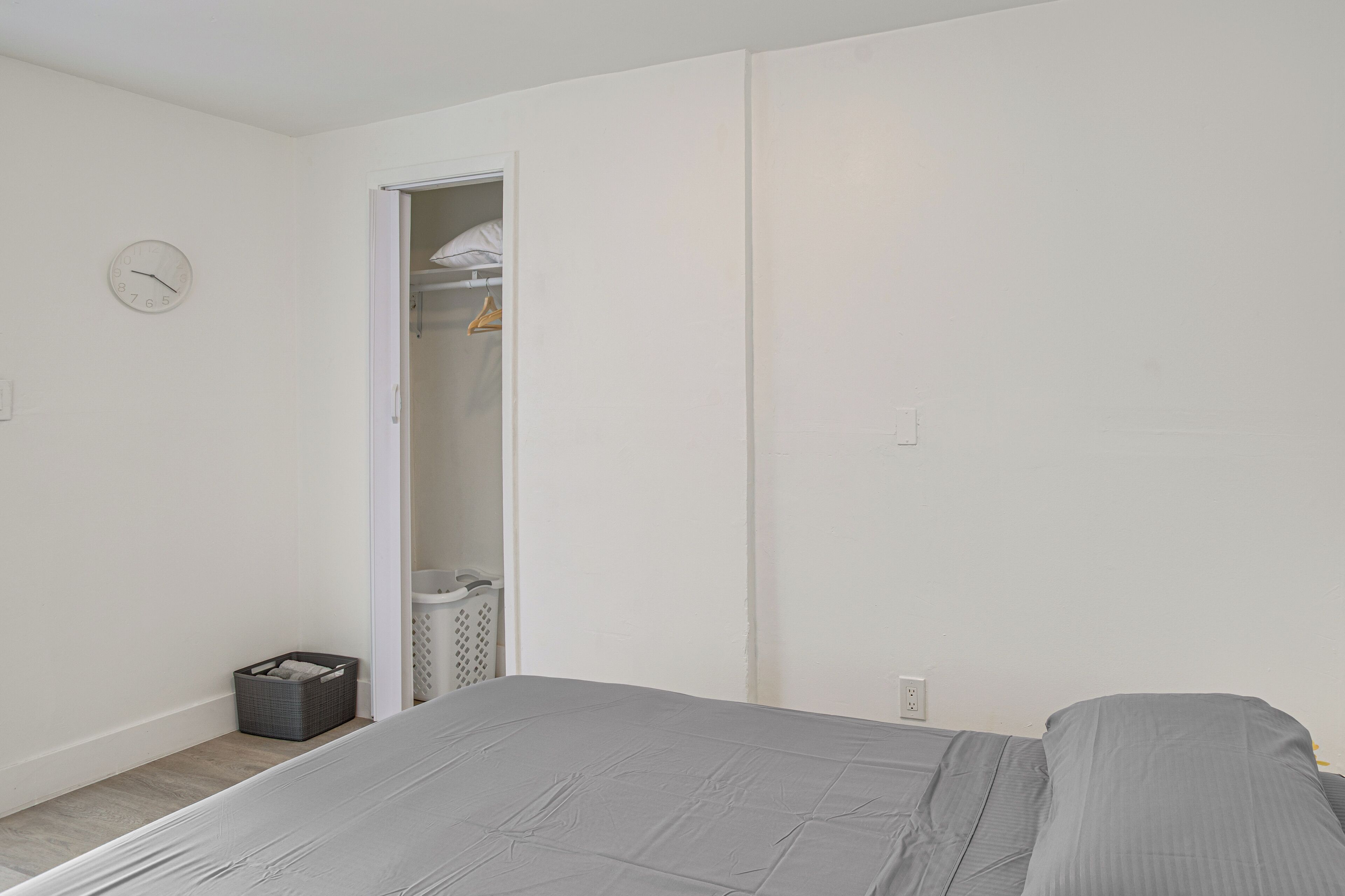 Ein Schlafzimmer mit Bett, Kleiderschrank mit Kleidung, Wäschekorb und einer Uhr an der Wand.