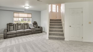 Une pièce recouverte de moquette, avec un canapé, une fenêtre équipée de stores, un escalier et une porte.