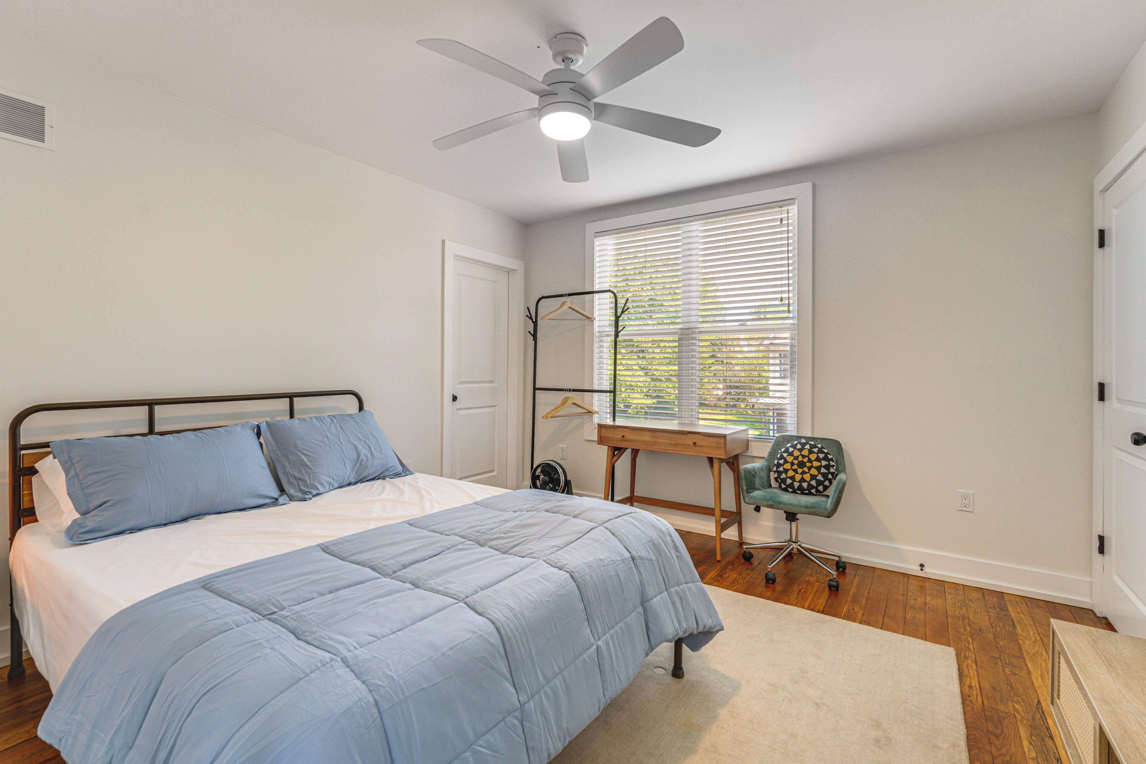 A bedroom with a bed, a desk, a chair, a ceiling fan, and a window with blinds.